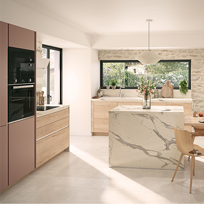la cuisine Dolce naturelle d’ixina une grande cuisine claire, avec un grand îlot central. L’îlot, tout en longueur, est divisé en une moitié plan de travail en marbre, et une autre moitié étant une table en bois claire autour de laquelle sont installées 4 chaises. Des assiettes sont sur cette table pour illustrer que l’on peut s’y attabler pour un repas.